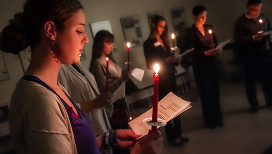 Aids vigil with a group of people standing in a circle in a dark room holding red candles and pieces of paper 