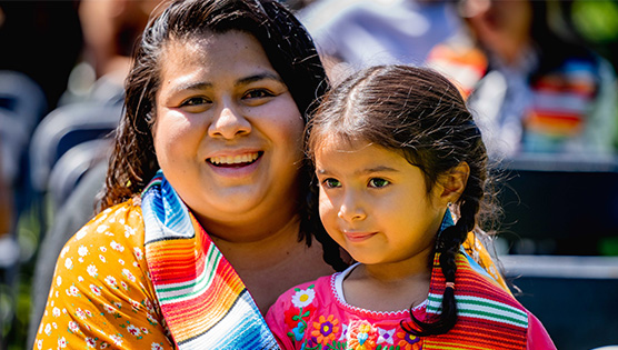 A graduating student who wears an orange shirt is smiling and holding her daughter in her lap while  with her serape inspired graduate stole is wrapped around them both