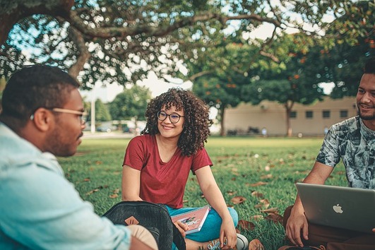 Three non-traditional students sitting on grass under a tree, talking and smiling, with a notebook and laptop nearby.