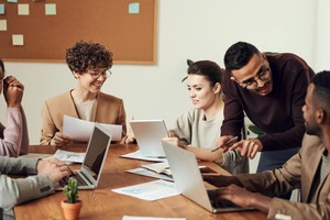 A group of diverse professionals collaborating around a table with laptops, papers, and a tablet. They are smiling and discussing ideas in a modern office setting.